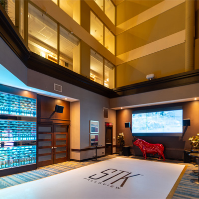 Modern hotel atrium lounge with wood tables, tan chairs, a large screen showing football, and contemporary wall fireplaces.