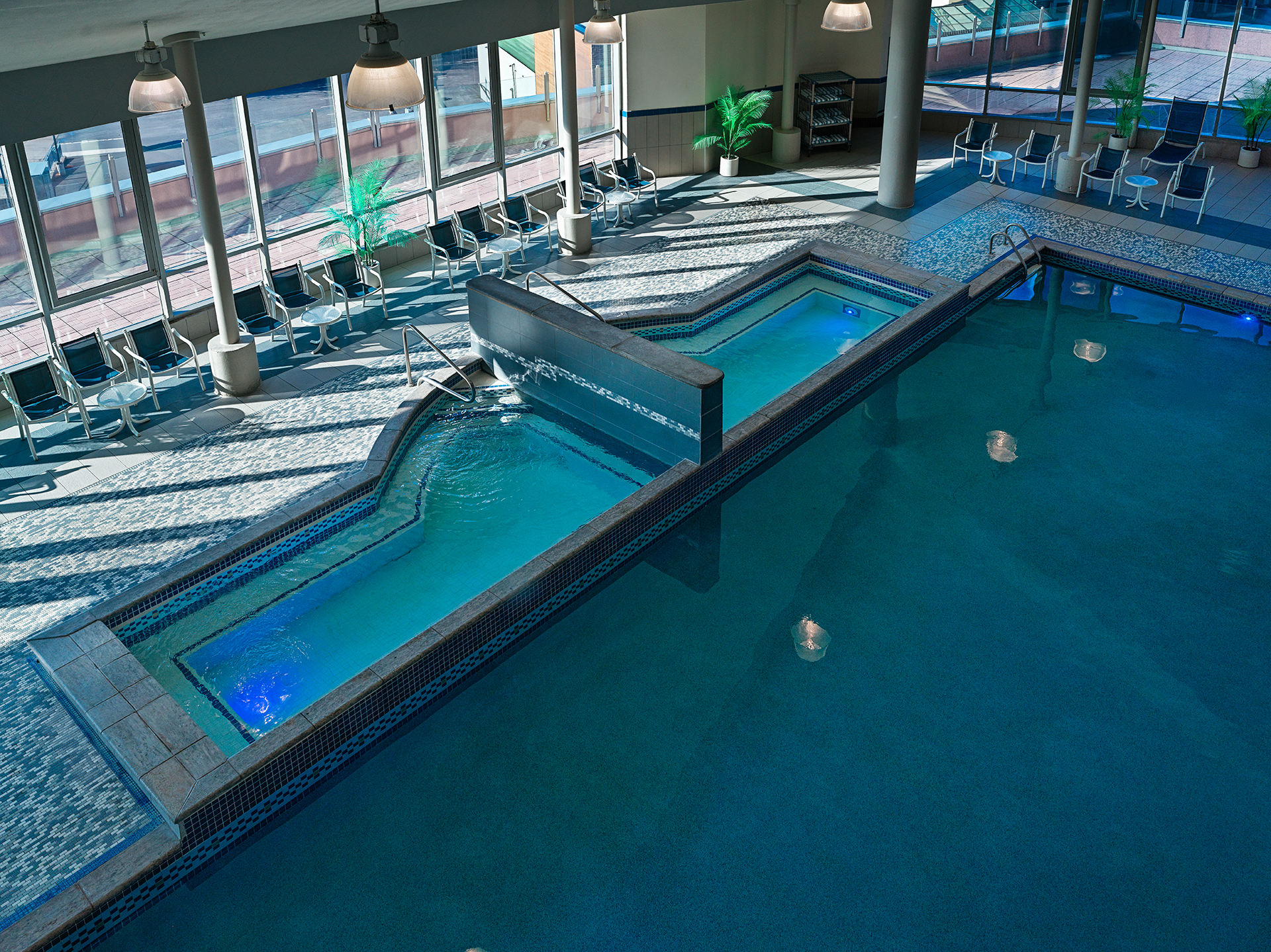 A large indoor swimming pool, with two hot tubs, large windows surrounding the pool deck with tables and lounge chairs.